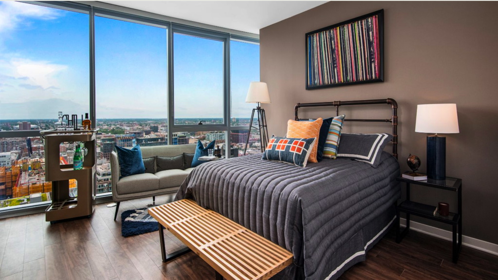 Modern bedroom with large windows, city view, gray bedding, wooden bench, sofa, bar cart, and abstract art on the wall.