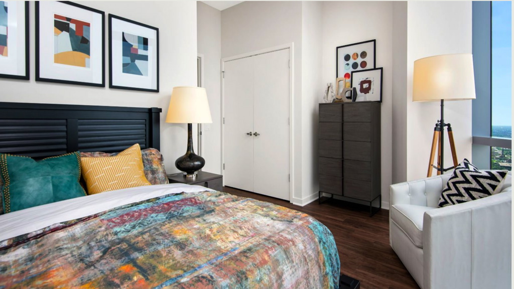 Modern bedroom with a large bed, colorful abstract artwork above, and a cushioned chair beside a tall lamp. A dresser with decor and a window offering city views complete the room.