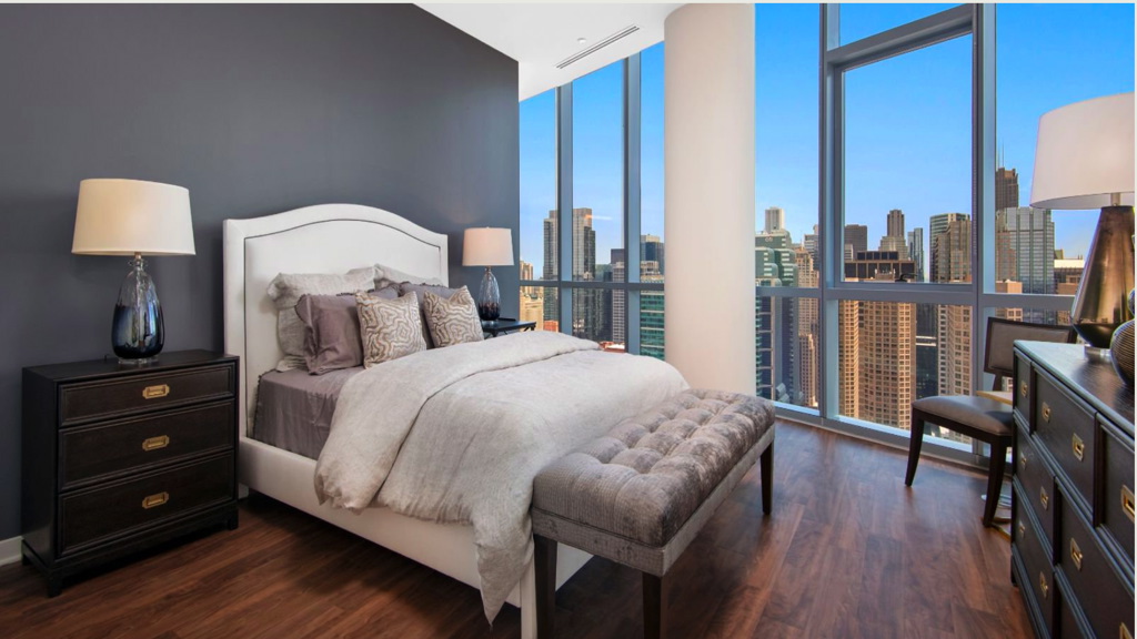 Modern bedroom with a large bed, gray walls, and cityscape view through floor-to-ceiling windows. Furniture includes two bedside tables with lamps and a cushioned bench at the foot of the bed.