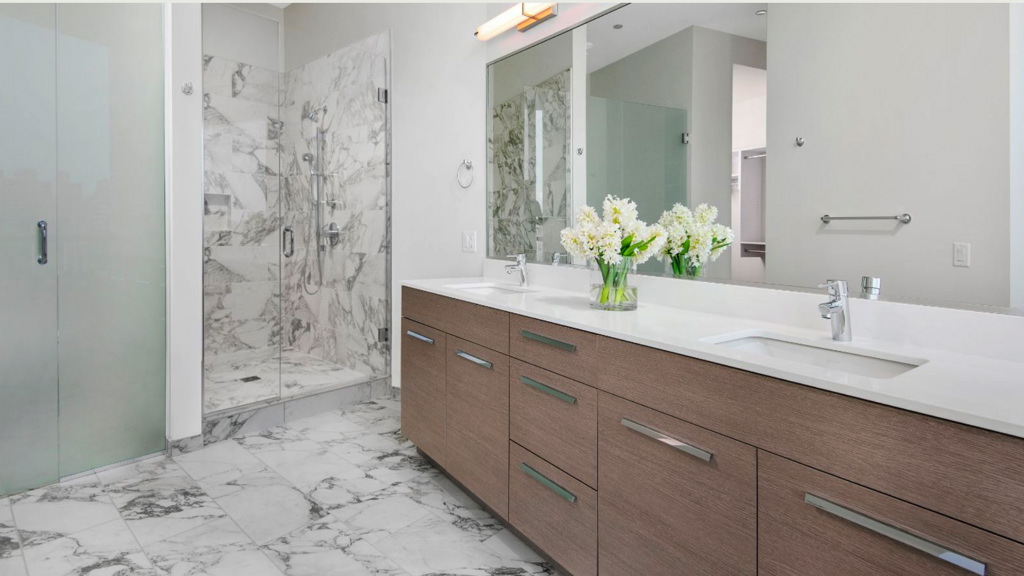 Modern bathroom with a glass-enclosed marble shower, a double sink vanity with wooden cabinets, and a vase of flowers. Walls and floor are light-toned, with ample natural light.