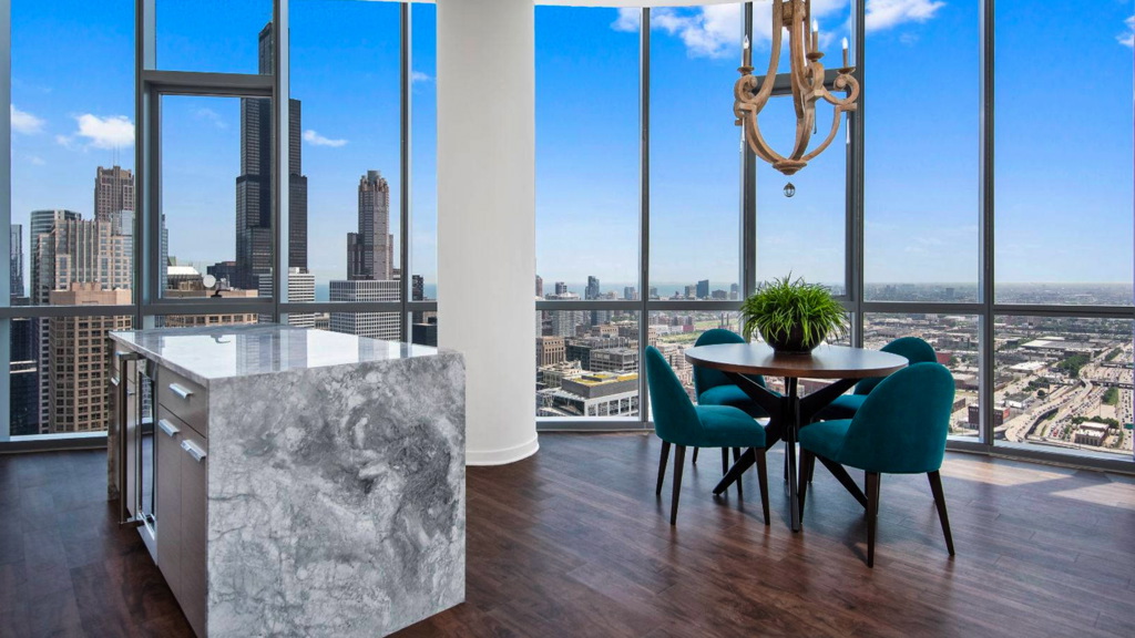 Modern high-rise apartment interior with floor-to-ceiling windows, city skyline view, marble kitchen island, teal dining chairs, and wooden floor.