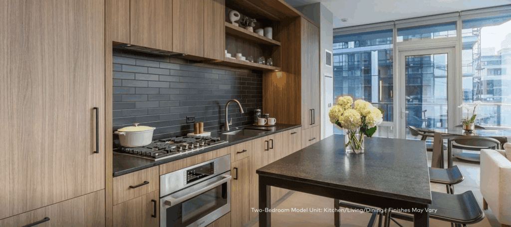 Modern kitchen with wood cabinetry, dark backsplash, built-in oven, gas stove, and a dining island with a vase of flowers; large windows show a cityscape view.