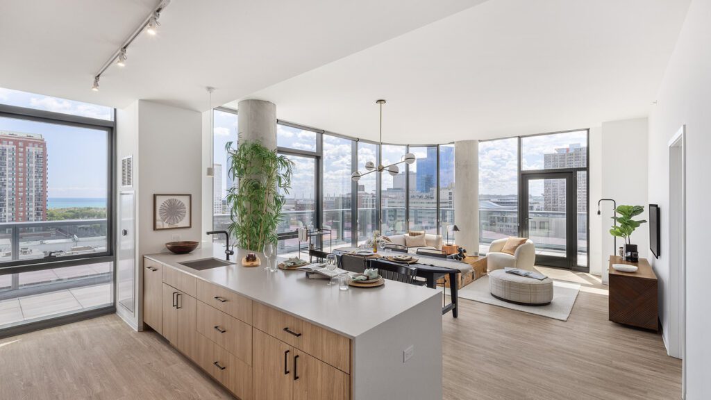 Modern open-plan apartment with large windows, kitchen island, dining area, and living space; cityscape and water visible outside.