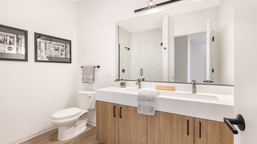 Modern bathroom with a double sink vanity, wood cabinets, white countertop, large mirror, toilet, wall art, and a glass-enclosed shower in the background.