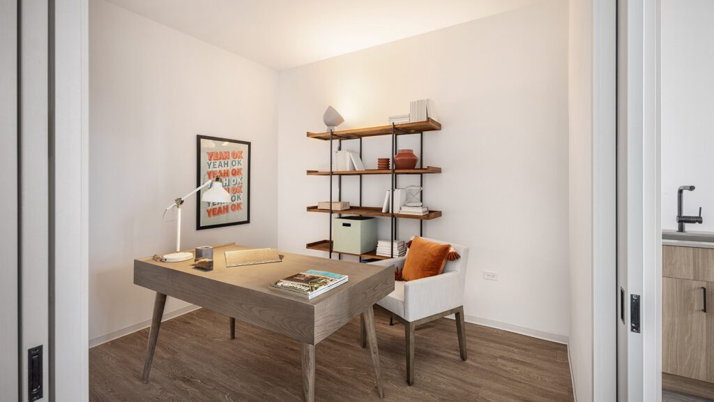 A modern home office with a wooden desk, a white chair with an orange pillow, a wall shelf with books and decor, and a framed print on a white wall.