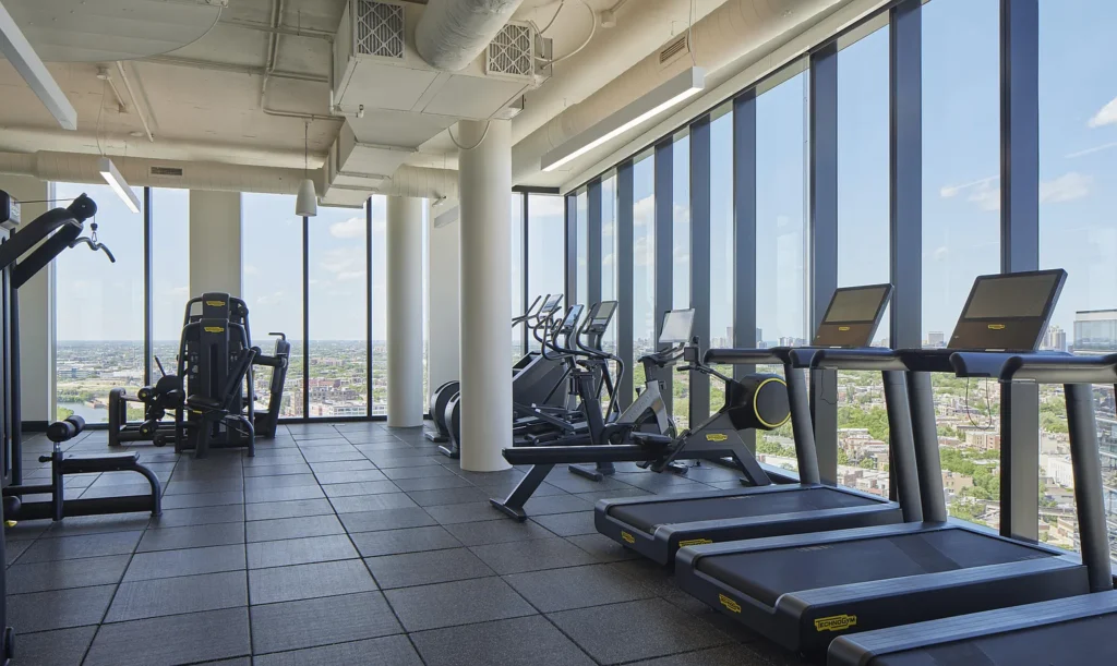 Modern gym with treadmills, elliptical machines, and strength equipment, featuring large floor-to-ceiling windows overlooking a cityscape.