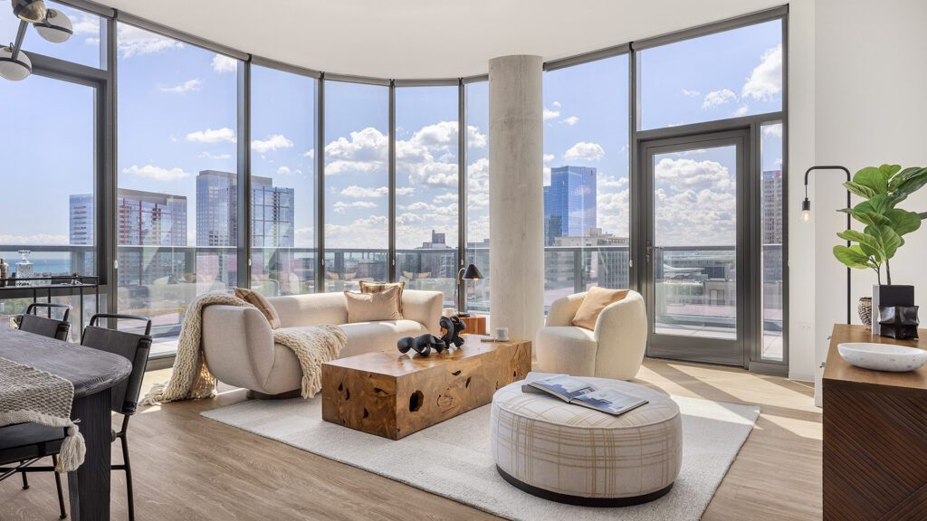 Modern living room with floor-to-ceiling windows, city views, neutral furniture, a wooden coffee table, and indoor plants in a bright, open space.