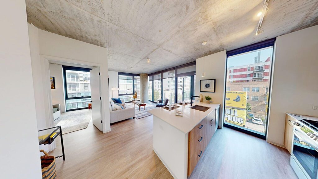 Modern apartment with open kitchen, wooden floors, large windows, and city views; living area and bedroom visible in bright natural light.