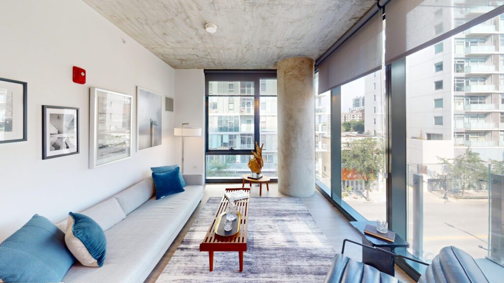 Modern living room with floor-to-ceiling windows, gray sofa, blue accents, coffee table, abstract art, and city views. Concrete ceiling and pillar visible.