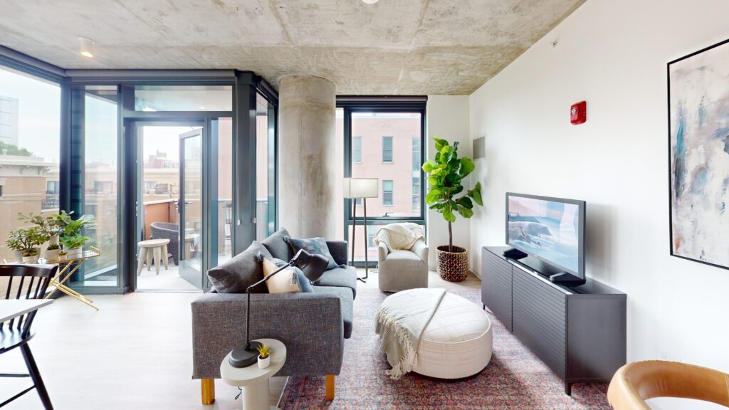 Modern living room with large windows, gray sofa, round ottoman, TV, potted plant, balcony access, and city view. Concrete ceiling and contemporary decor.
