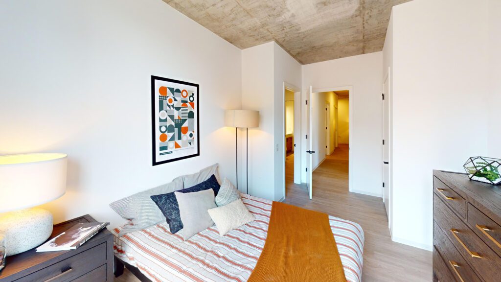 A bedroom with a striped bed, orange blanket, abstract wall art, floor lamp, dresser, and exposed concrete ceiling. A hallway and two doors are visible in the background.