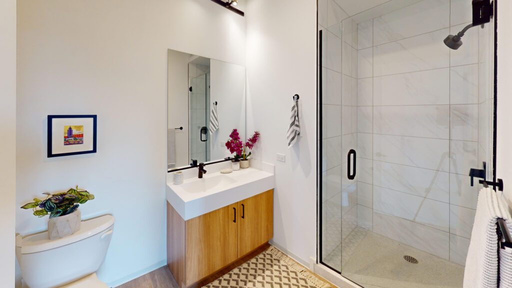 Modern bathroom with a wooden vanity, white sink, large mirror, glass shower, toilet, wall art, and vase with flowers on the counter.