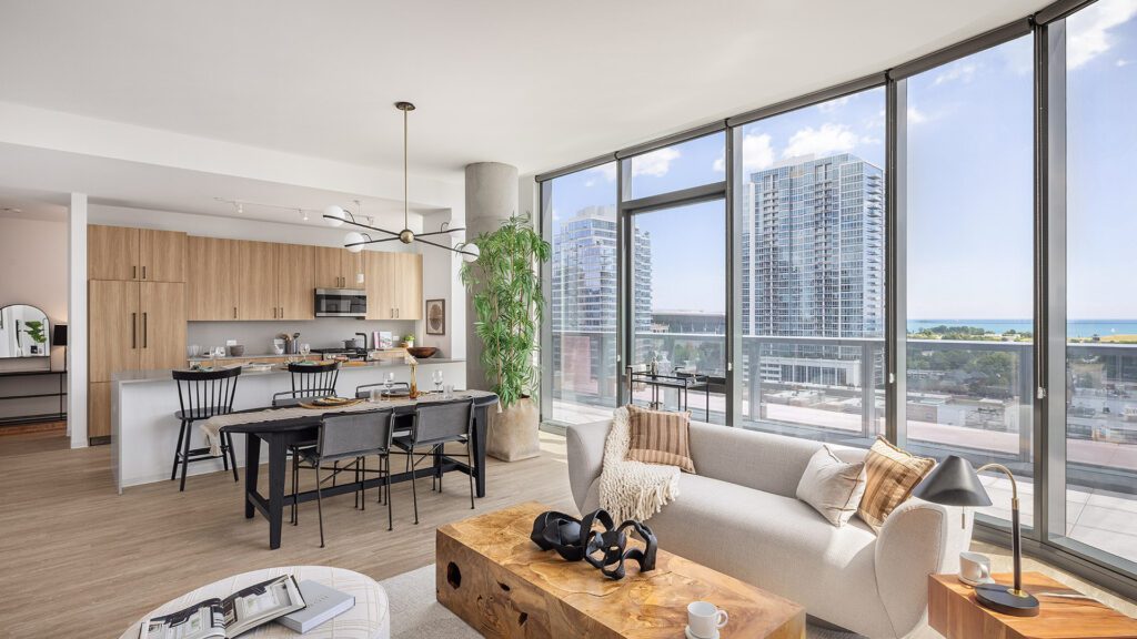 Modern open-concept apartment with large floor-to-ceiling windows, light wood flooring, a kitchen with bar seating, dining area, and living room overlooking city buildings and water.