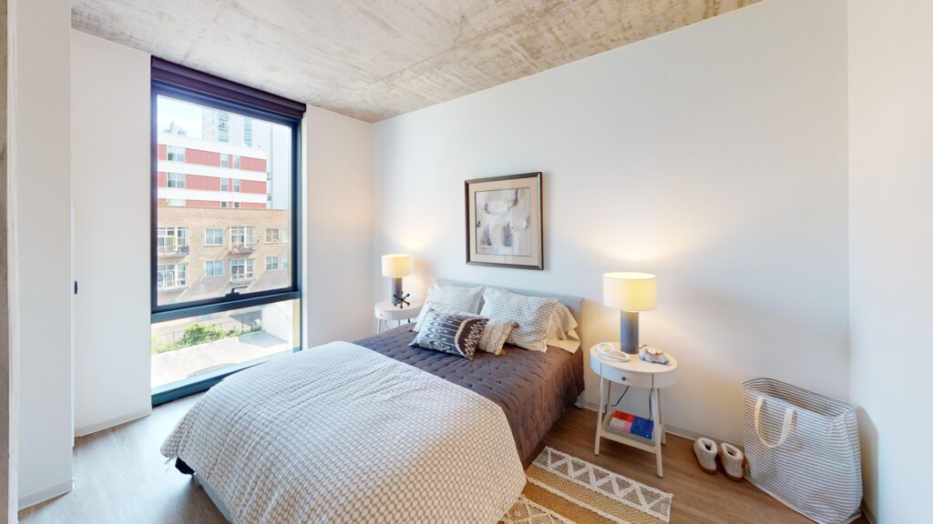 A modern bedroom with a large window, a neatly made bed with pillows and blankets, two bedside tables with lamps, and a decorative basket on the floor.
