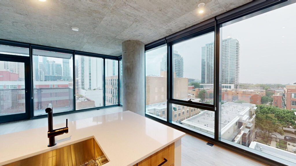 Modern apartment interior with large floor-to-ceiling windows, a kitchen island with a sink in the foreground, and a cityscape view with high-rise buildings in the background.