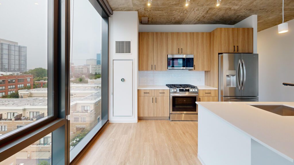 Modern kitchen with wood cabinets, stainless steel appliances, white countertops, and large windows overlooking a cityscape.