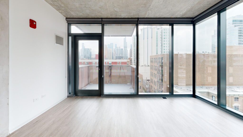 Unfurnished modern apartment room with floor-to-ceiling windows, a glass door leading to a balcony, and city buildings visible outside.