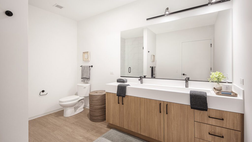 Modern bathroom with a double sink vanity, large mirror, toilet, towel rack, wicker laundry basket, and light wood cabinets. Neutral tones and minimal decor.
