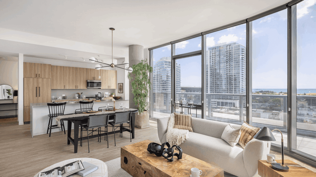 Modern open-concept apartment with large floor-to-ceiling windows, light wood flooring, a kitchen with an island, dining area, and living space overlooking city buildings and a glimpse of water.
