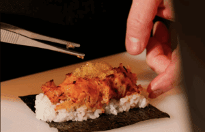 A person uses tweezers to place garnish on top of a sushi roll made with rice, grilled topping, and seaweed.