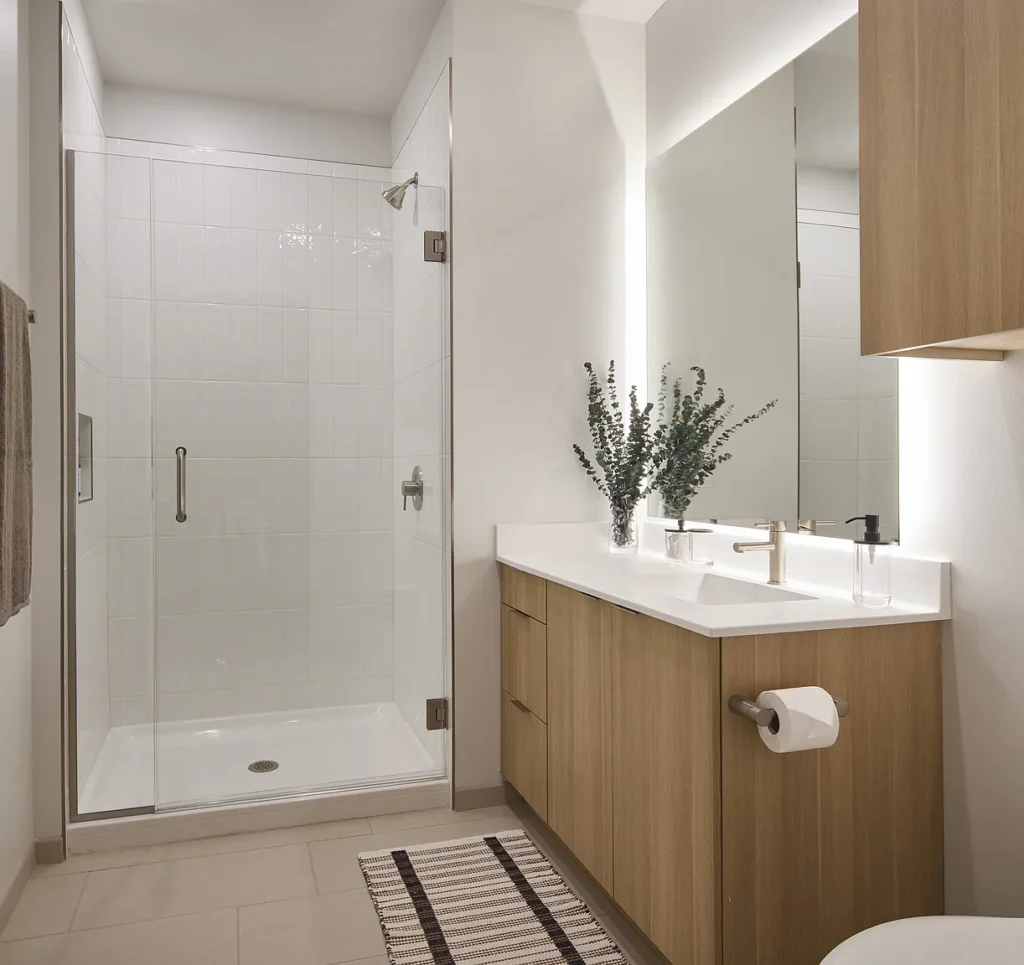 Modern bathroom with a glass shower enclosure, wood vanity with sink, large mirror, wall-mounted faucet, and a striped rug on the tiled floor.