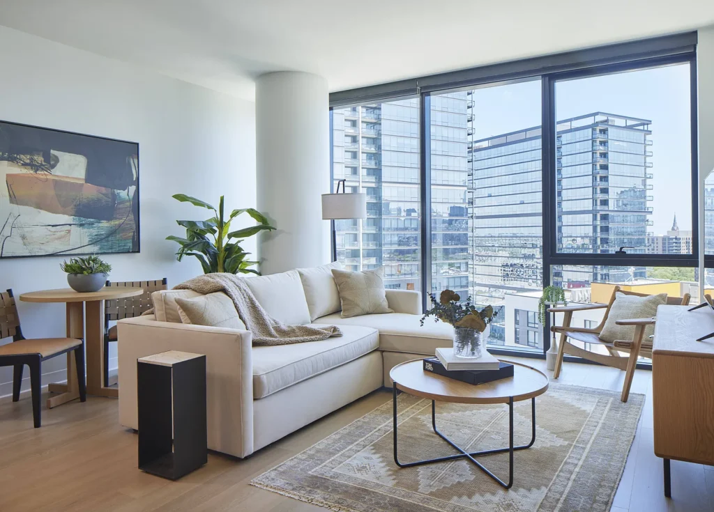 A modern living room with a white sofa, round coffee table, indoor plants, large windows, and cityscape views, featuring neutral tones and contemporary decor.