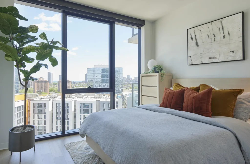 A modern bedroom with large floor-to-ceiling windows, a city view, a neatly made bed with brown pillows, a potted plant, dresser, and abstract wall art.