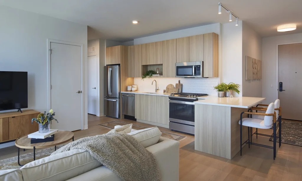 Modern apartment kitchen and living area with light wood cabinets, stainless steel appliances, a small island with stools, and a sofa facing a TV.