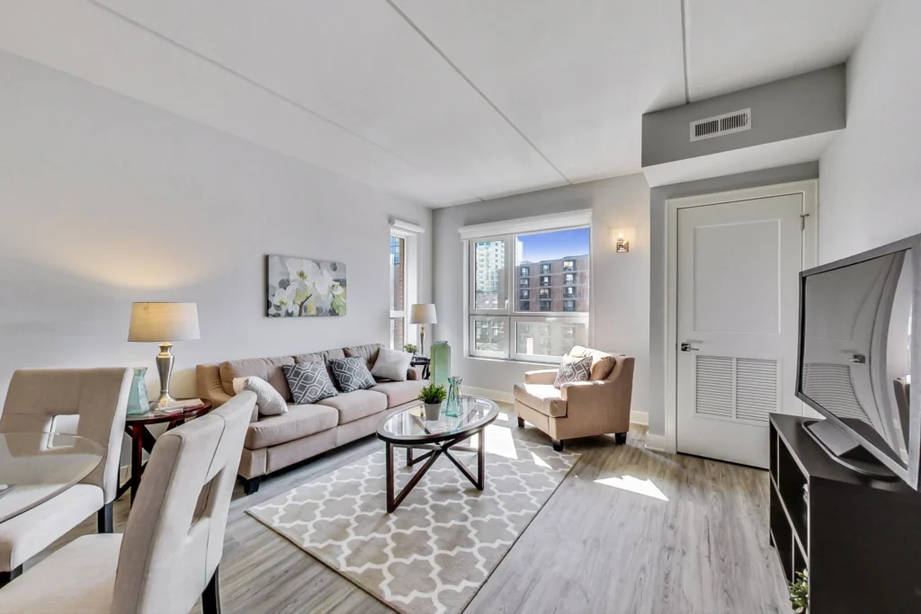 Bright living room with a beige sofa, armchair, glass coffee table, TV, dining area, large window, and neutral decor.