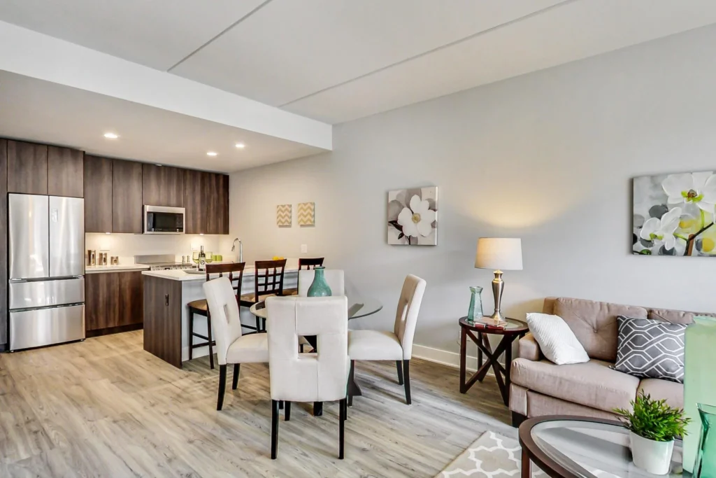 Modern open-plan kitchen and living room with wood flooring, stainless steel appliances, dining table, beige sofa, and floral wall art.