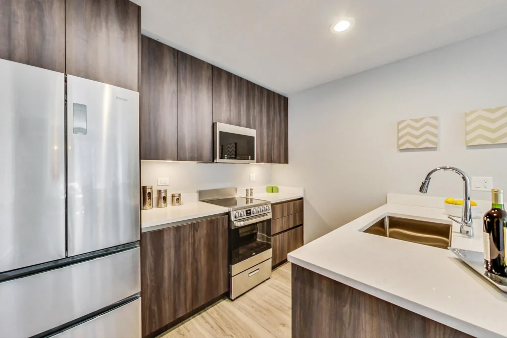 Modern kitchen with stainless steel appliances, wood cabinets, a white countertop, a sink, and decorative wall art. A bottle of wine and lemon slices are on the counter.