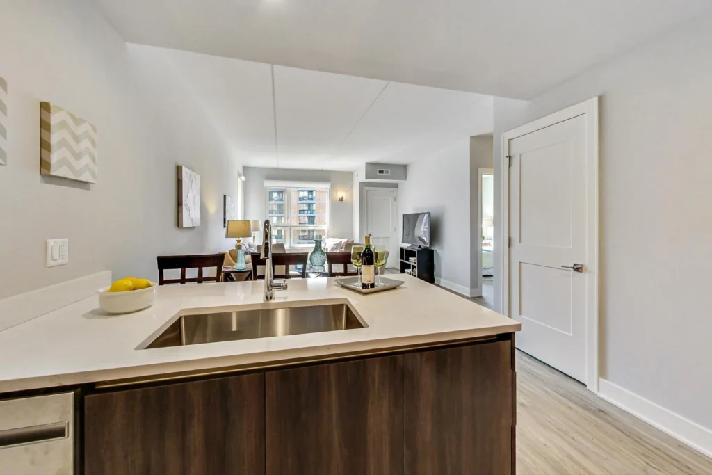 Modern apartment interior showing a kitchen island with sink in the foreground, and a living room with seating and TV in the background.