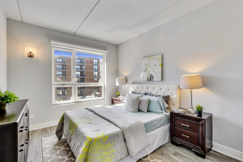 A modern bedroom with a large window, double bed, two bedside tables with lamps, a dresser, and wall art above the headboard.