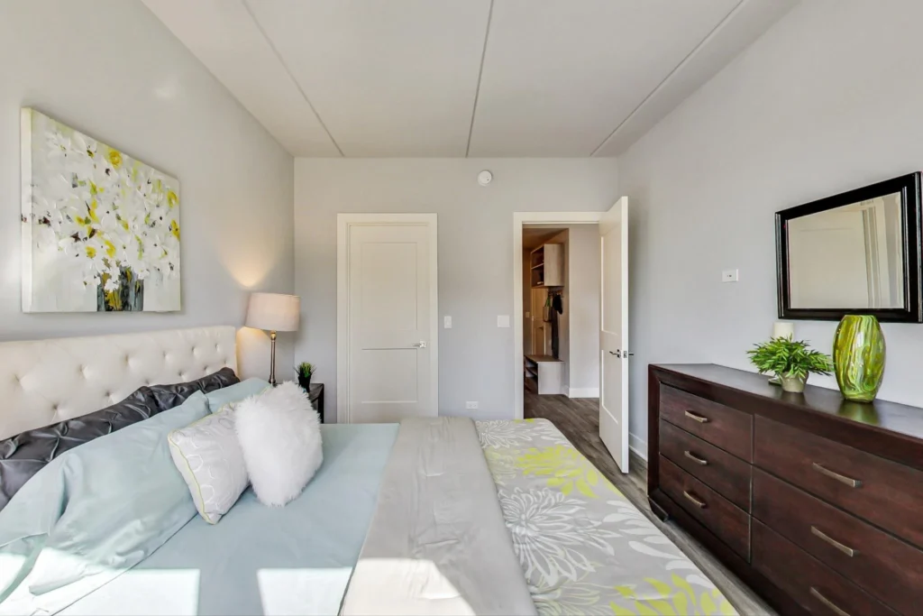 A neatly made bed with patterned bedding is beside a nightstand and lamp; a dresser with plants and a mirror is against the opposite wall in a bright bedroom.