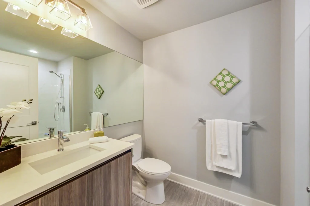 Modern bathroom with a large mirror, wooden vanity, white countertop, toilet, towel rack with white towels, and a green wall accent. Shower with glass door is visible in the background.