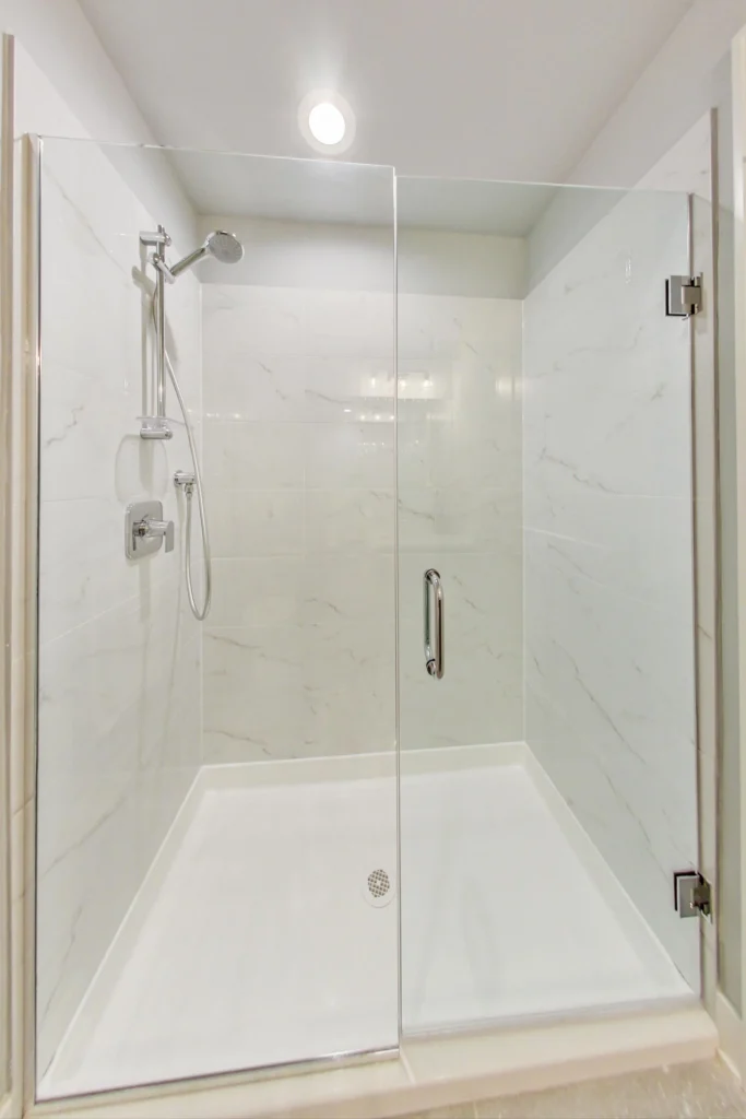 A modern glass-enclosed shower with a handheld showerhead, wall tiles with a marble pattern, and a metal handle on the door.