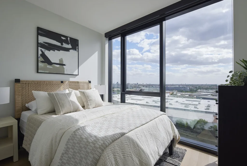 Modern bedroom with a large bed, neutral decor, abstract wall art, and floor-to-ceiling windows overlooking an urban landscape under a partly cloudy sky.
