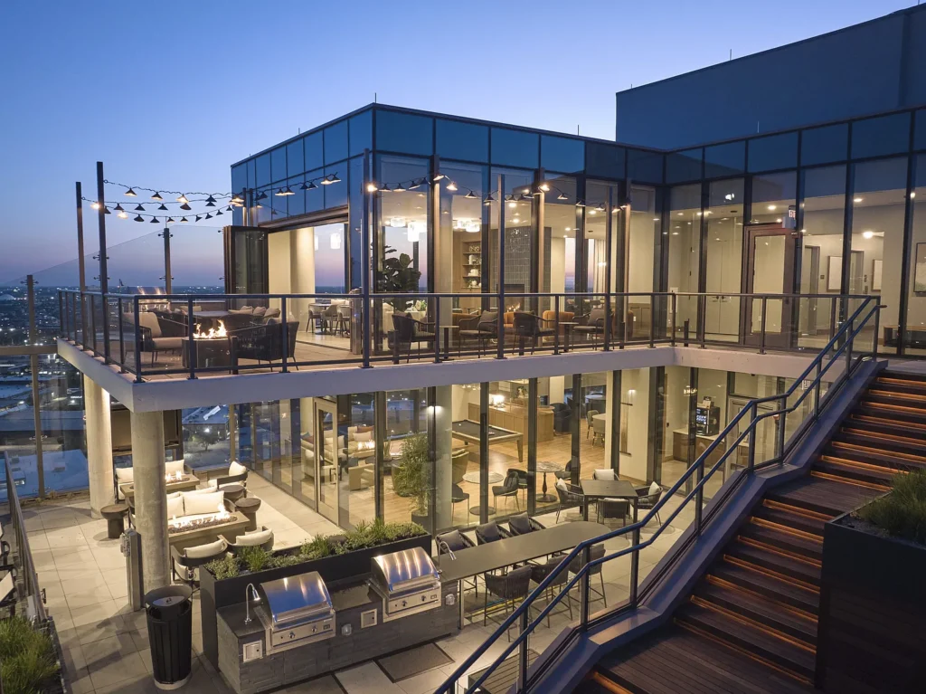 Modern rooftop terrace at sunset with glass-walled lounge, outdoor seating, fire pit, string lights, and built-in barbecue grills. Stairs with lit steps lead to the upper level.