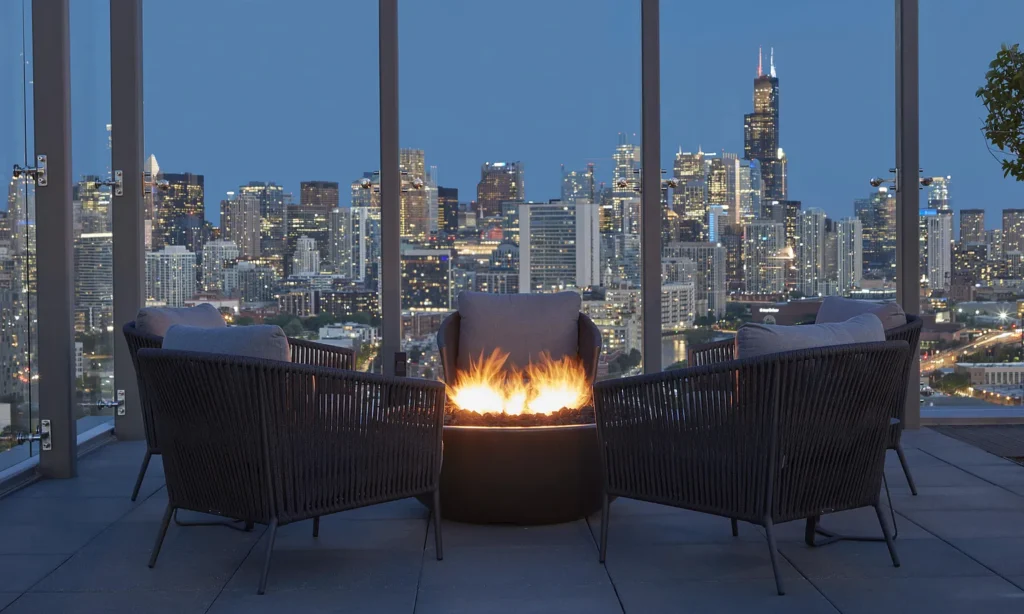 Three chairs surround a fire pit on a balcony at night, overlooking a city skyline with illuminated skyscrapers.