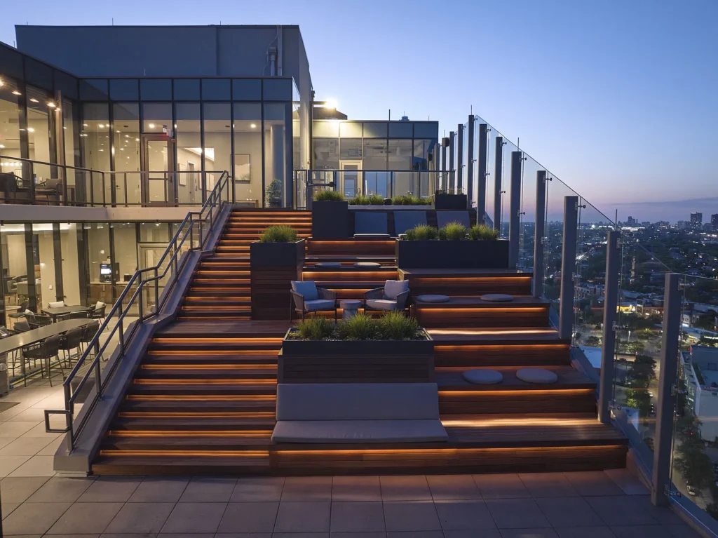 Modern rooftop terrace at dusk with tiered wooden seating, glass railings, built-in planters, and city views in the background.