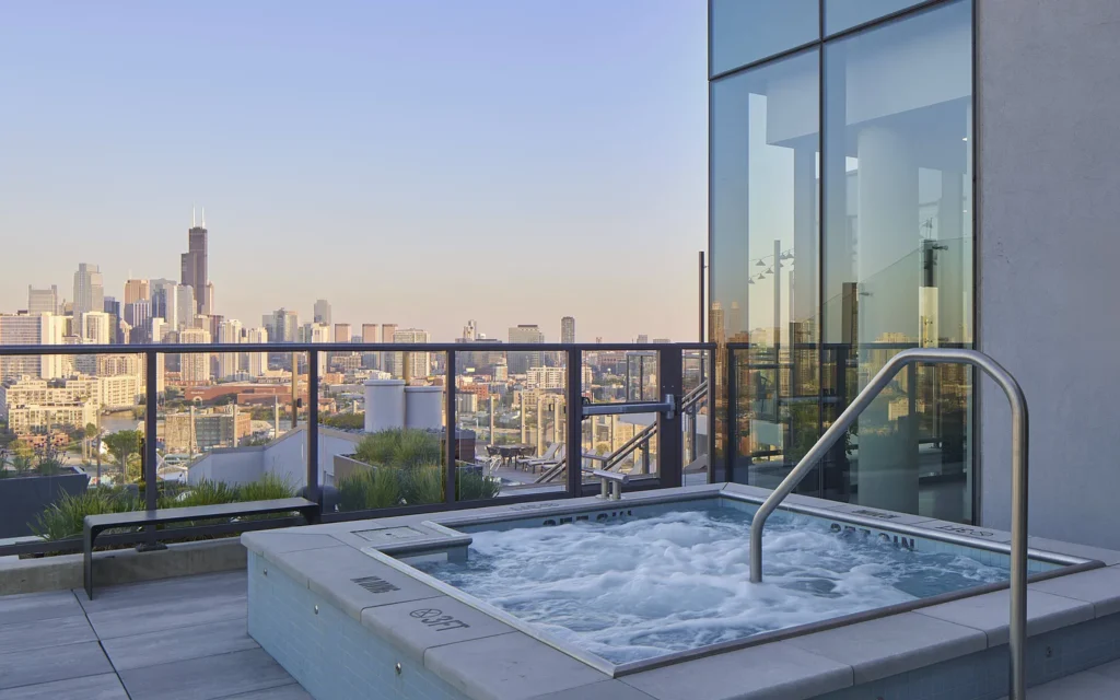 A rooftop hot tub with bubbling water overlooks a city skyline at sunset, with glass railings and a modern building nearby.