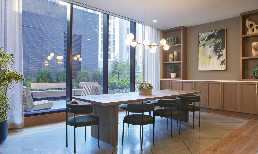 Modern dining room with a wooden table, six chairs, a contemporary chandelier, built-in cabinets, large windows, and a view of an outdoor patio with greenery.