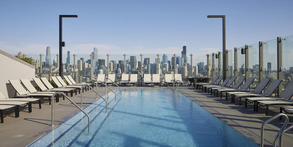 Rooftop swimming pool with lounge chairs on both sides, glass barriers, and a city skyline in the background under a clear blue sky.