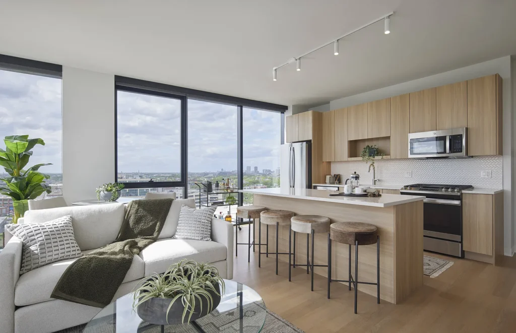 Modern apartment with large windows, open-plan kitchen with light wood cabinets, island seating, stainless steel appliances, and a living area with a white sofa and indoor plants.
