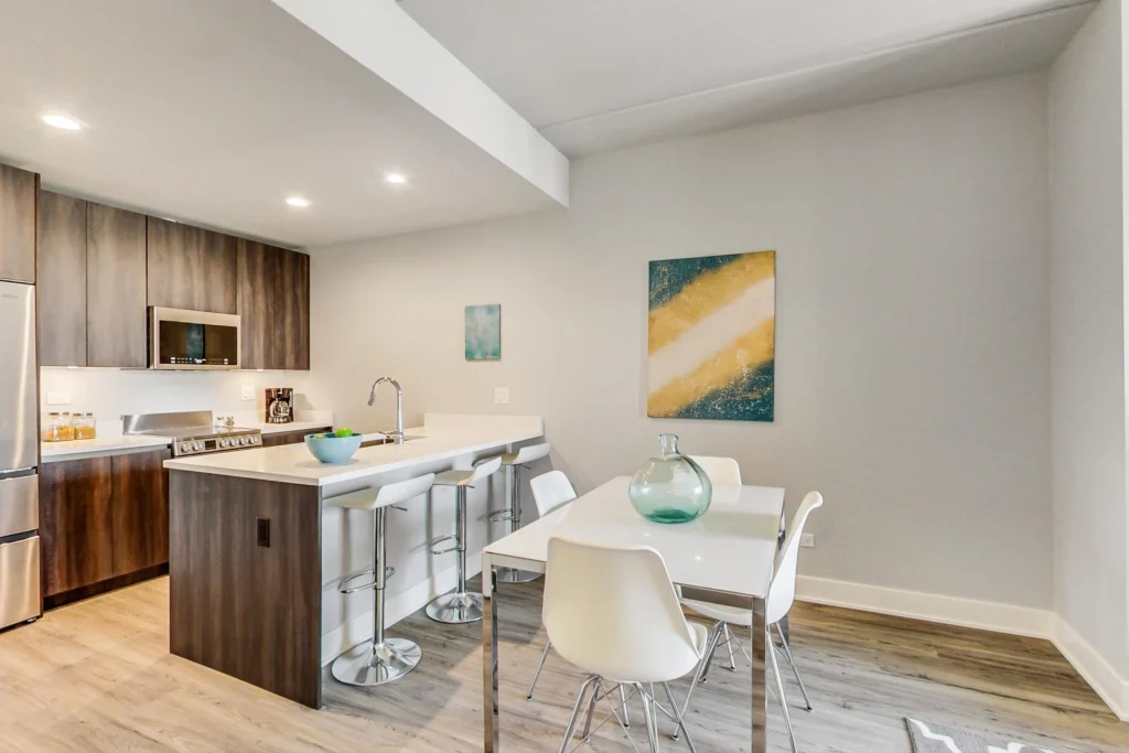 Modern kitchen and dining area with wood cabinets, stainless steel appliances, a white island with barstools, a table with four chairs, and abstract art on light gray walls.