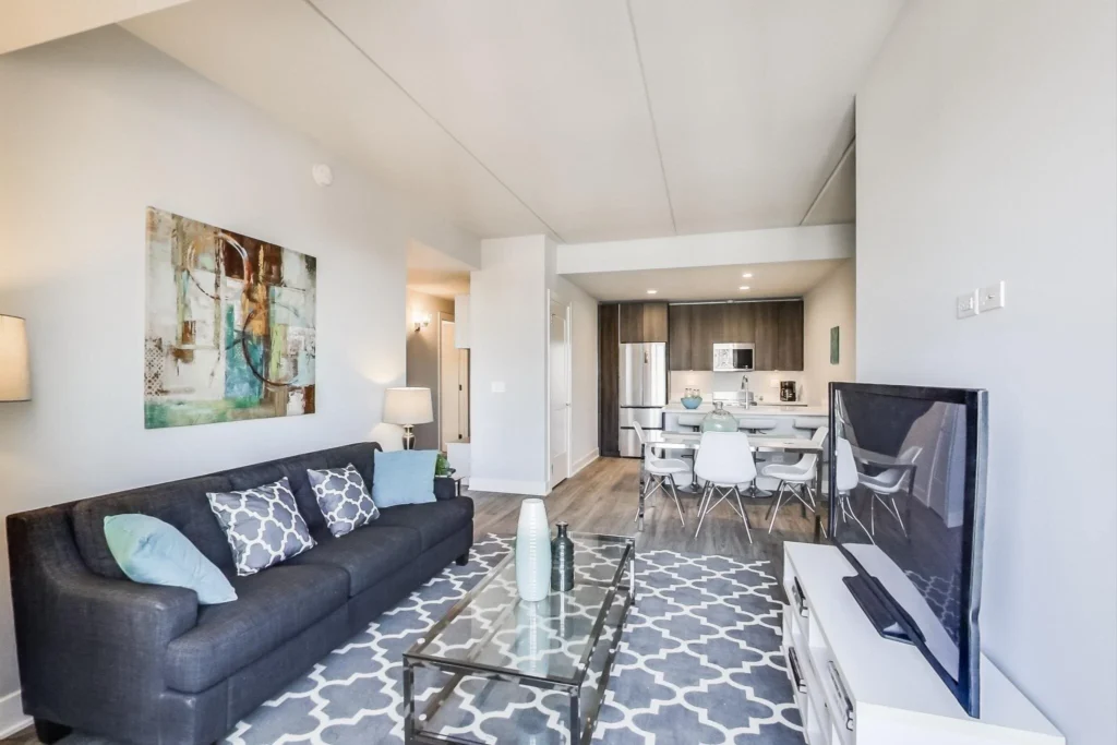 Modern open-concept living room and kitchen with gray sofa, patterned rug, glass coffee table, wall art, and dining area with white chairs. TV faces the seating area.