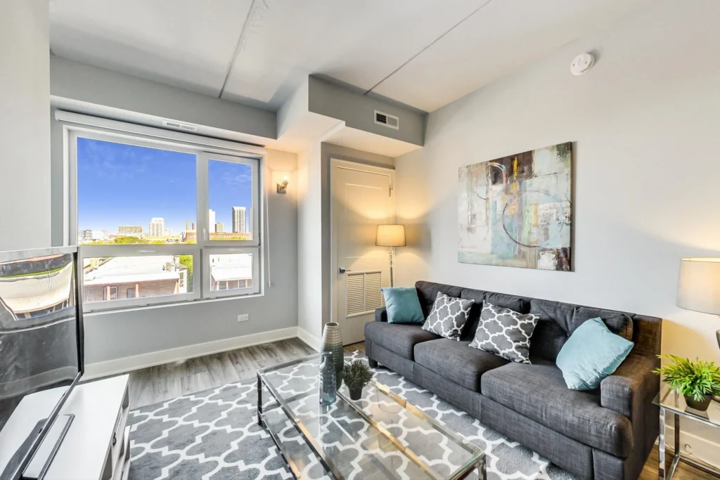 Modern living room with a gray sofa, patterned rug, glass coffee table, abstract wall art, and large window overlooking a cityscape.