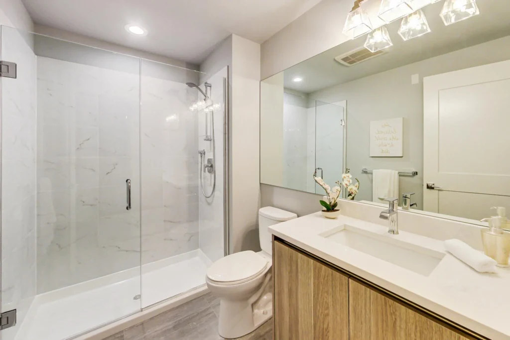 Modern bathroom with a glass-enclosed shower, toilet, large mirror, wooden vanity with white countertop, and orchid plant on the sink.