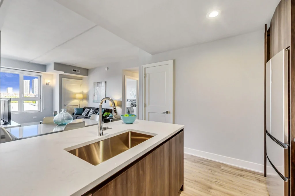 Modern kitchen with a white countertop island, stainless steel sink, wood cabinets, and view into a bright living area with large windows.
