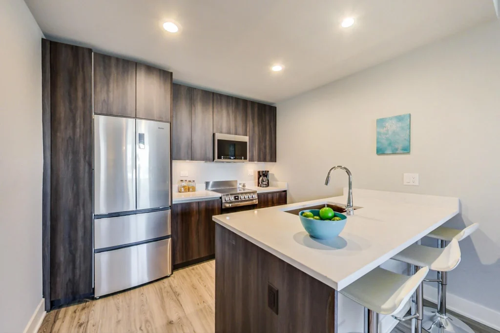Modern kitchen with stainless steel appliances, wood cabinets, a white countertop island with a bowl of fruit, and two white bar stools. A small painting hangs on the wall.